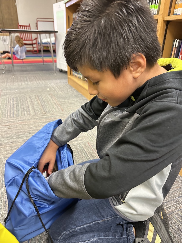 A young student kneels on the floor in a school library, reaching into a blue drawstring backpack. He is wearing a black and gray hoodie and jeans. Bookshelves and other students sitting on a colorful rug are visible in the background.