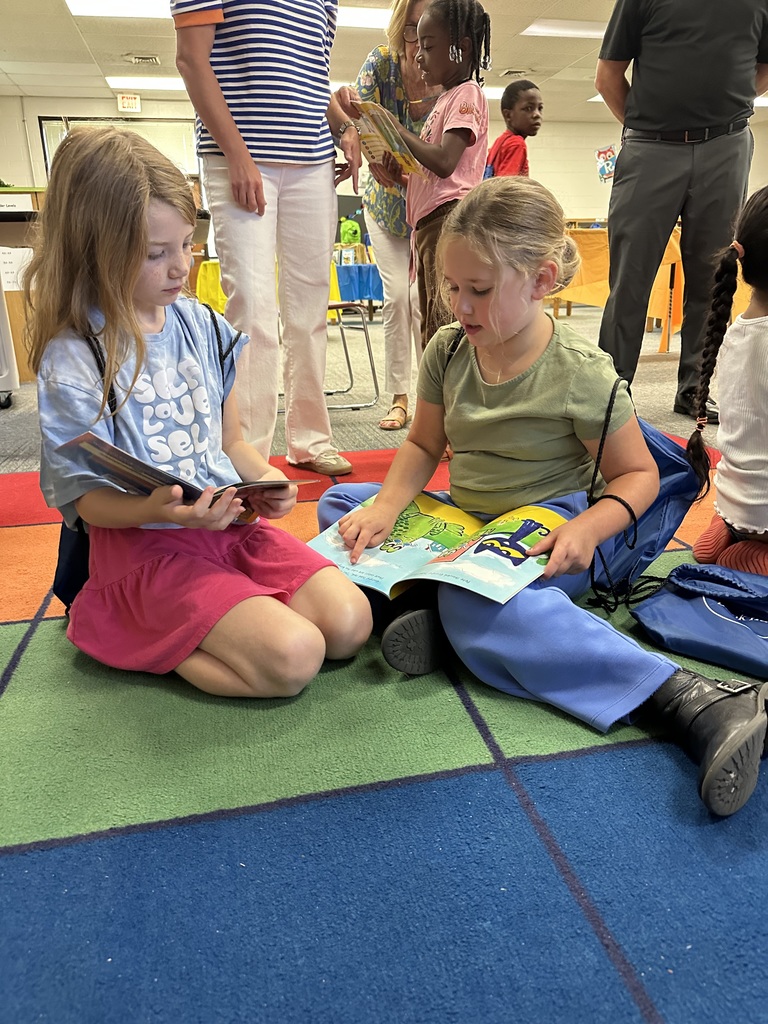 Two girls sit cross-legged on a colorful classroom rug, reading books together. One holds a Pete the Cat book while the other looks on. Adults and other students browse books in the background.