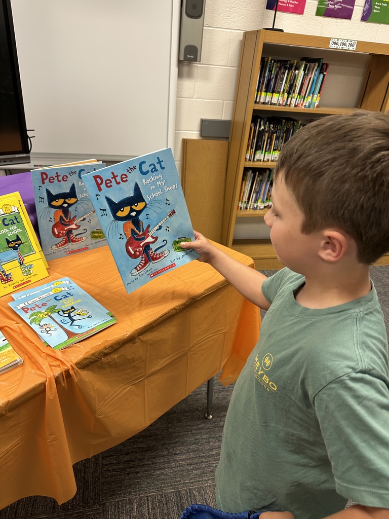 A young boy stands in a school library holding a copy of Pete the Cat: Rocking in My School Shoes while looking at a display of Pete the Cat books arranged on an orange-covered table. Shelves of books and a whiteboard are visible in the background.
