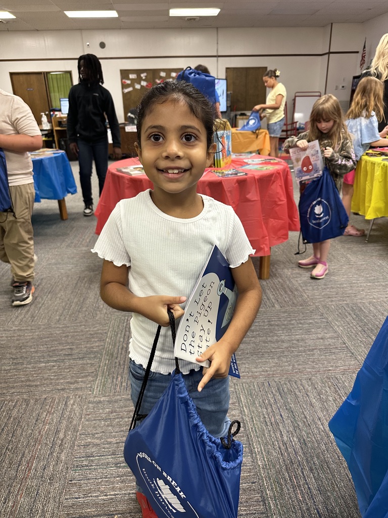 A smiling young girl stands in a library holding a copy of Don't Let the Pigeon Stay Up Late! while gripping a blue "Books on Break" drawstring bag. Other children are visible in the background browsing books.