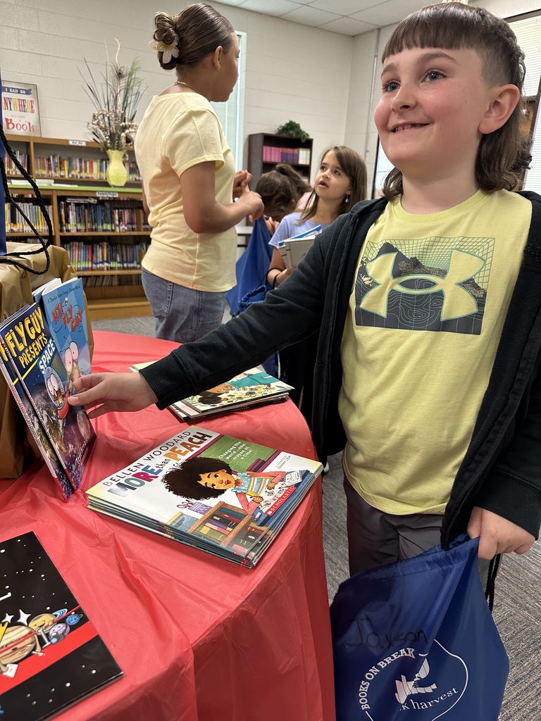 A smiling boy stands at a table covered with a red tablecloth, reaching for a book while holding his blue "Books on Break" bag. The table displays a variety of colorful children’s books. Other students browse in the background.