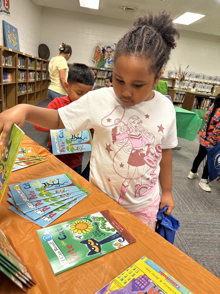 A young girl wearing a Barbie-themed shirt browses a table full of colorful children's books, including several titles from the Pete the Cat series. She holds a blue drawstring book bag in one hand while reaching for a book with the other. Other students and shelves of books are visible in the background of the school library.