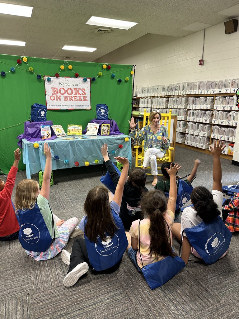 A group of elementary students wearing blue "Books on Break" drawstring backpacks sit on the floor, attentively listening to a woman seated in a yellow rocking chair. Behind her is a colorful display featuring a green backdrop, a sign that reads “Welcome to Books on Break,” and a table filled with children’s books. The setting appears to be a school hallway or media center decorated for the event.