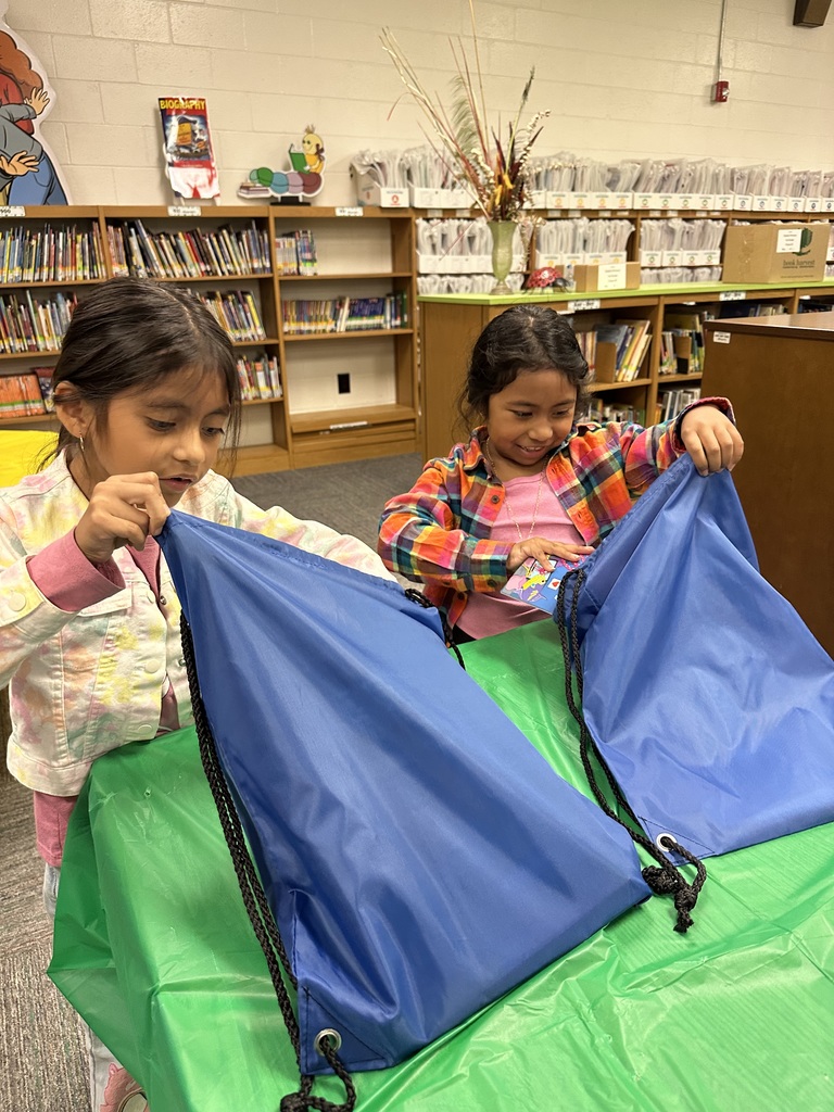 Two young girls stand at a green-covered table in a school library, excitedly opening their blue drawstring book bags. Shelves filled with books and labeled bins line the background, along with colorful decorations and a tall vase of dried floral arrangements.