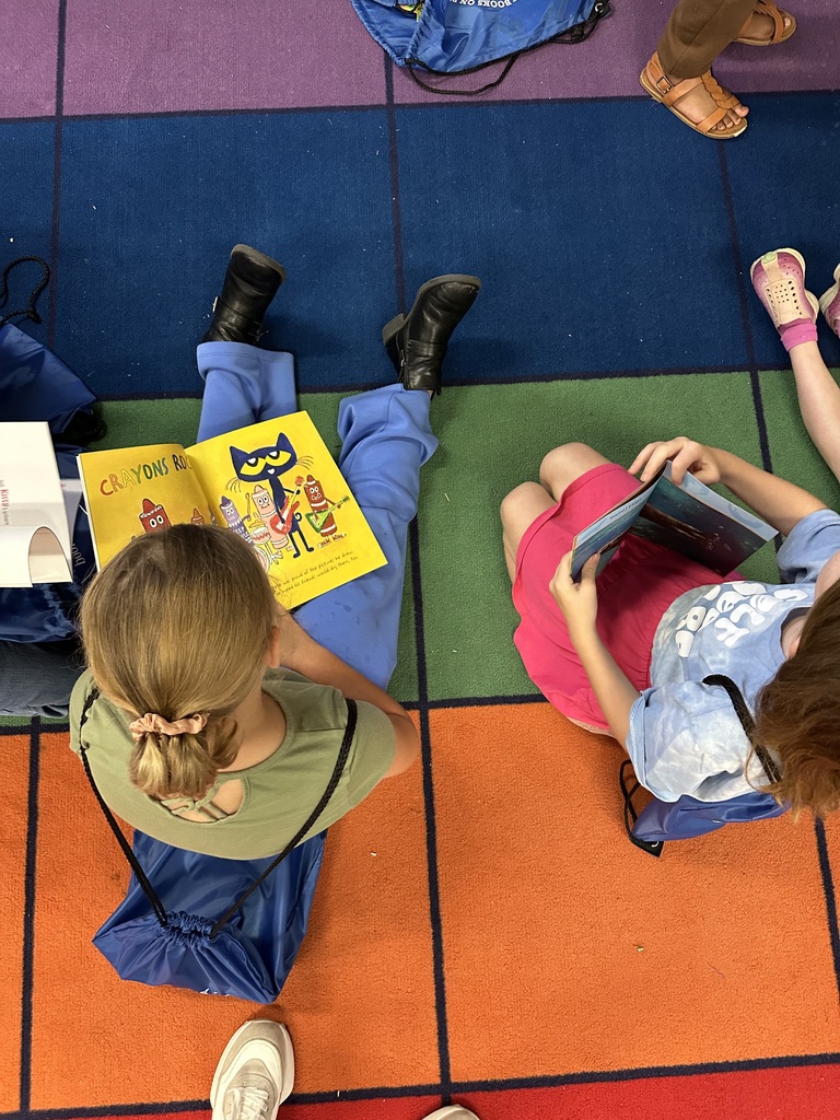 Top-down view of two girls sitting on a colorful rug reading books. One girl is reading Crayons Rock! while both have their blue “Books on Break” drawstring bags nearby.