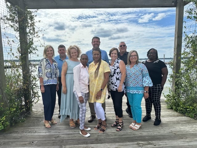 A group of ten adults stands together smiling on a wooden dock framed by greenery and a wooden overhead structure. Behind them is a scenic view of water and sky with light clouds. The group is casually dressed in colorful, summery attire, suggesting a relaxed outdoor gathering or professional retreat.