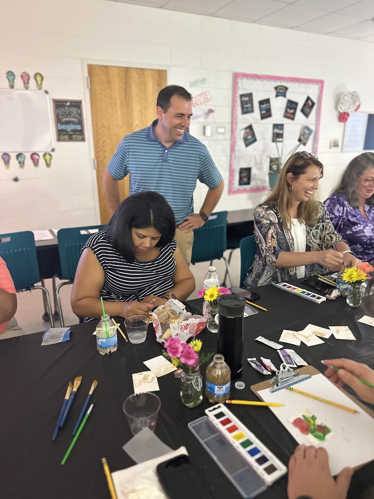 Man standing and smiling while watching women paint at a table.