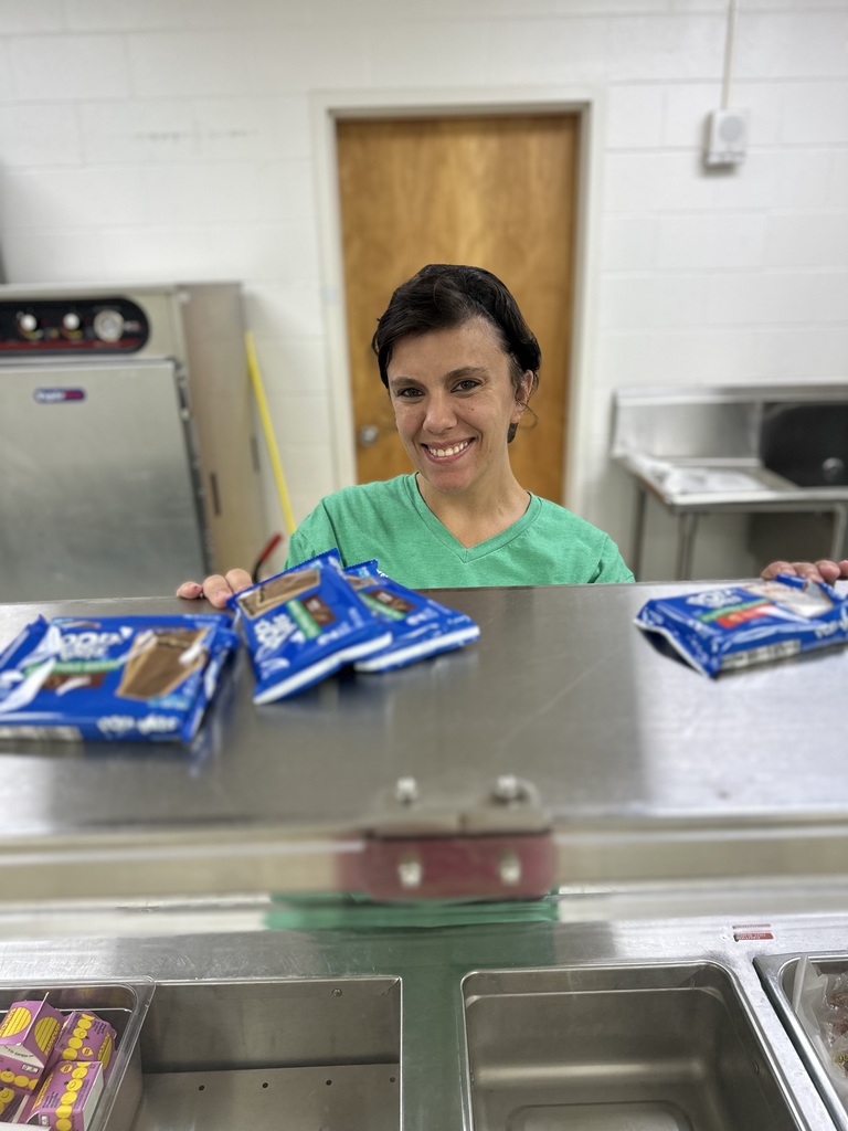 A cafeteria worker in a green shirt smiles from behind the serving counter with ice cream sandwiches on top.