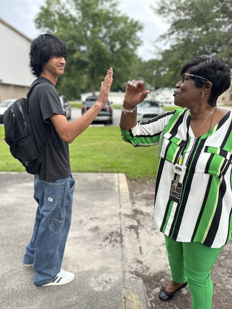 A student and a woman in a striped shirt and green pants exchange a high-five outside on a sidewalk