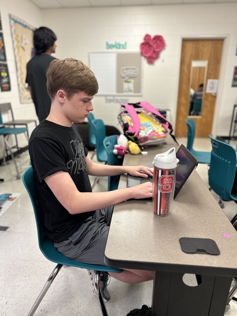 A student in a black T-shirt types on a laptop at a desk with a red-and-white tumbler; classroom items are visible in the background