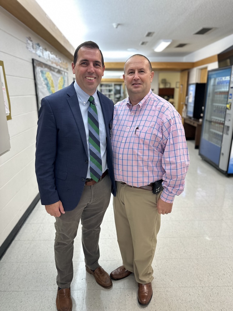 Two men stand side by side in a school hallway smiling at the camera; one wears a suit, the other a pink-and-blue plaid shirt.