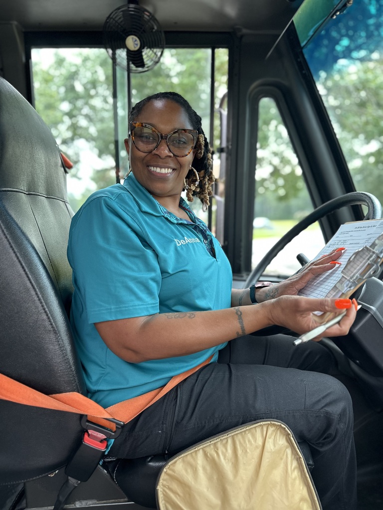 A school bus driver wearing glasses and a teal shirt smiles while holding a clipboard inside the bus.