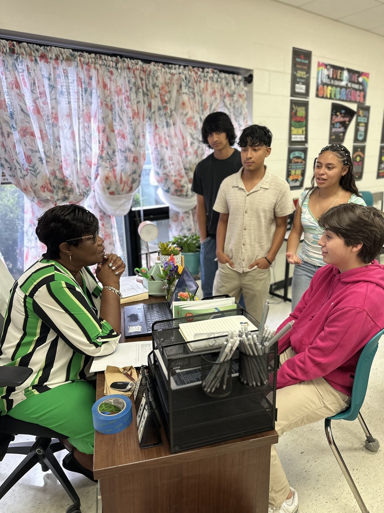 A woman in a striped shirt and green pants sits at a desk, talking with four standing students in a classroom