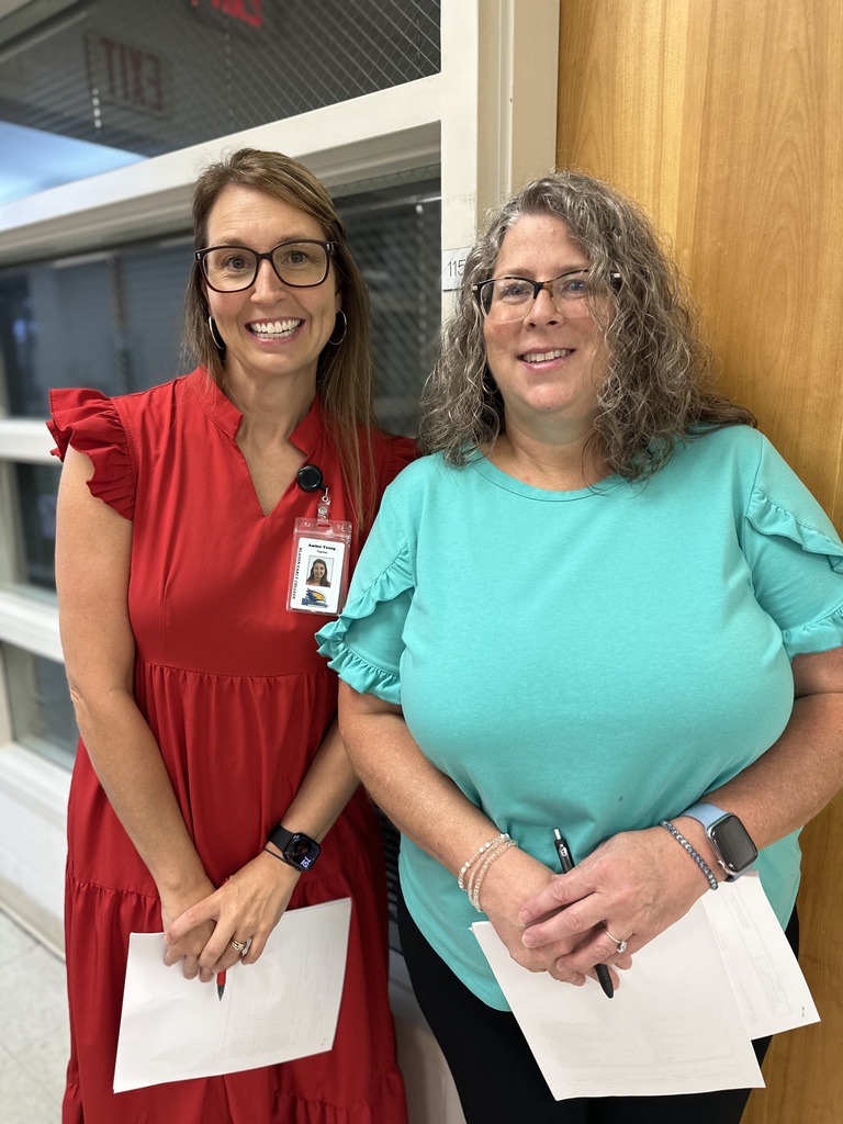 Two smiling women stand indoors holding papers; one wears a red dress and glasses, the other a teal top and glasses.