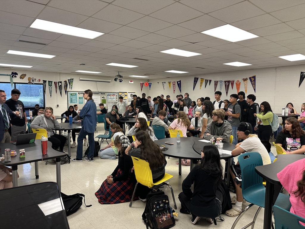 A large group of students sit and stand in a classroom, listening to a speaker at the front; college pennants line the walls