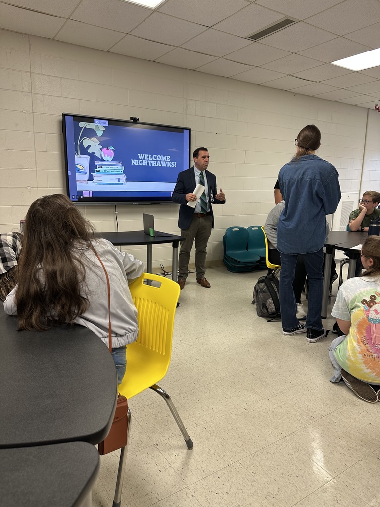 A man in a suit speaks to students in a classroom with a screen displaying “Welcome Nighthawks!” behind him