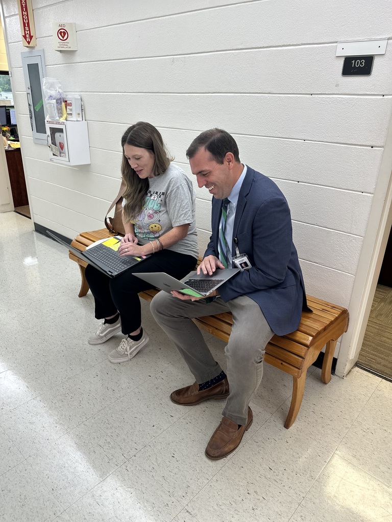 A man in a suit and a woman in a graphic T-shirt sit on a bench in a hallway, working on laptops together