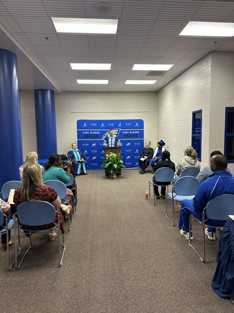 A wide shot of a graduation ceremony with attendees seated facing a man in academic regalia speaking at a podium in front of an East Bladen Eagles backdrop.