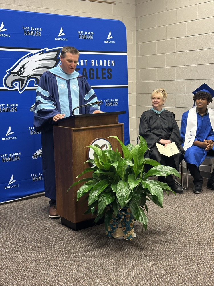 A man in academic regalia speaks at a podium in front of an East Bladen Eagles backdrop, with a woman in a black robe and a seated graduate in a blue cap and gown smiling beside him.