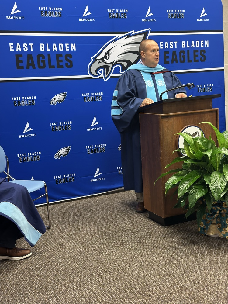 A man in academic regalia speaks at a podium in front of an East Bladen Eagles backdrop, with part of another person’s robe and shoe visible in the frame.