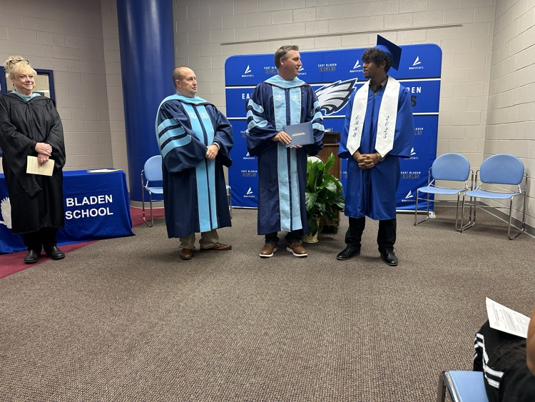 A graduate in a blue cap and gown stands on stage with two men in academic regalia, one handing him a diploma, while a woman in a black robe looks on.