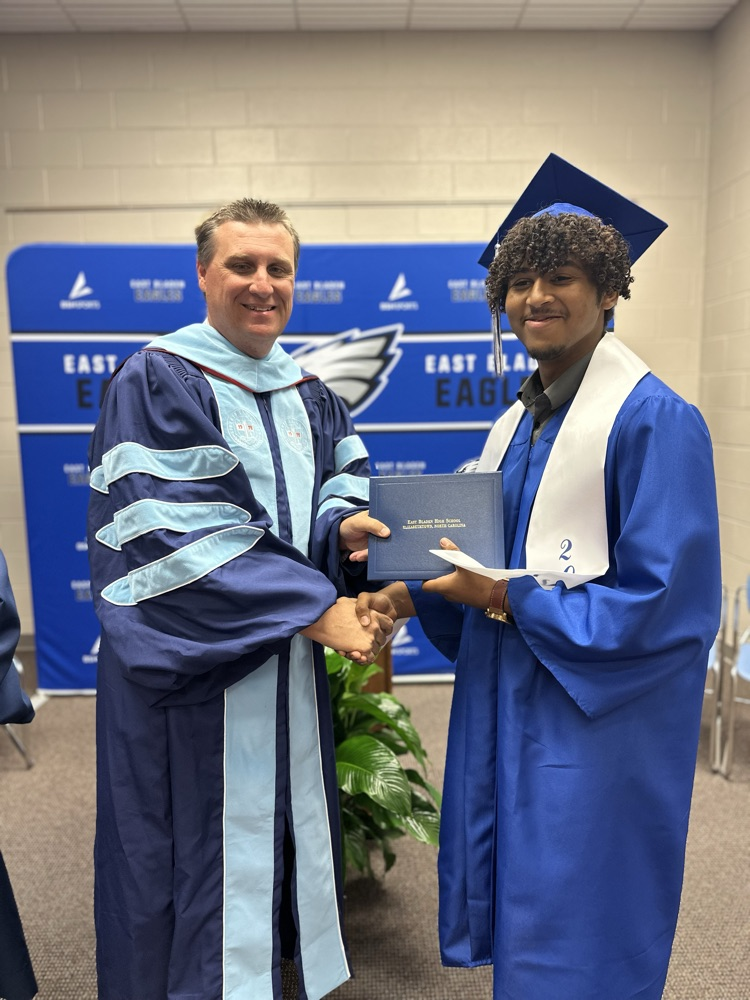 A graduate in a blue cap and gown with a white stole shakes hands with another man in academic regalia while holding a diploma in front of an East Bladen Eagles backdrop.