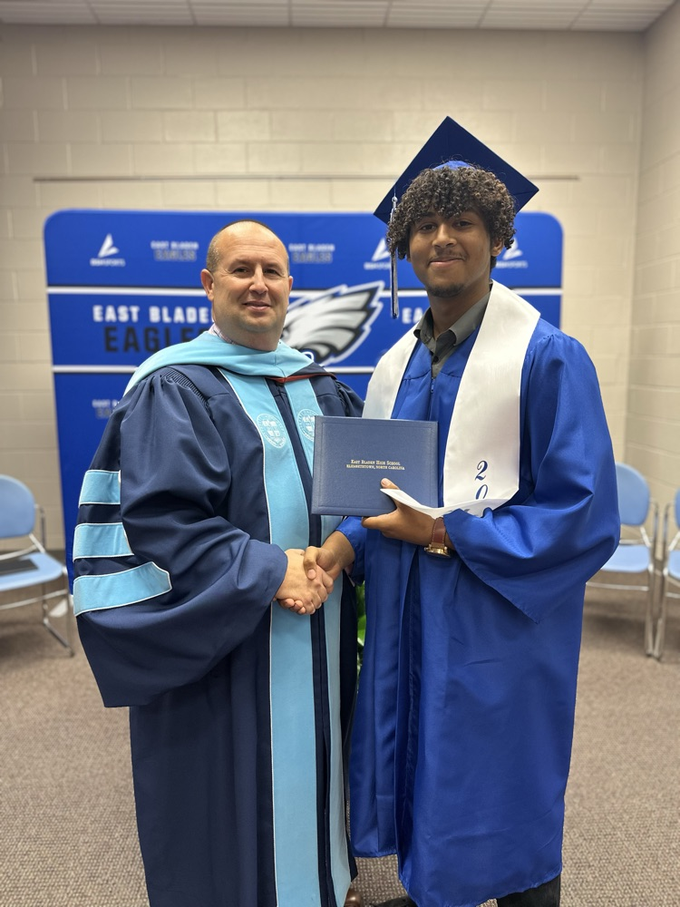 A graduate in a blue cap and gown with a white stole shakes hands with a man in academic regalia, both smiling at the camera while holding a diploma in front of an East Bladen Eagles backdrop.
