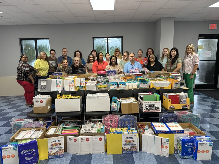 Group of people standing behind tables filled with donated school supplies for students and classrooms.
