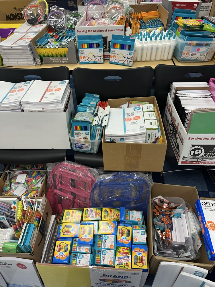 Boxes of school supplies including paper, glue, markers, crayons, backpacks, and other classroom items.