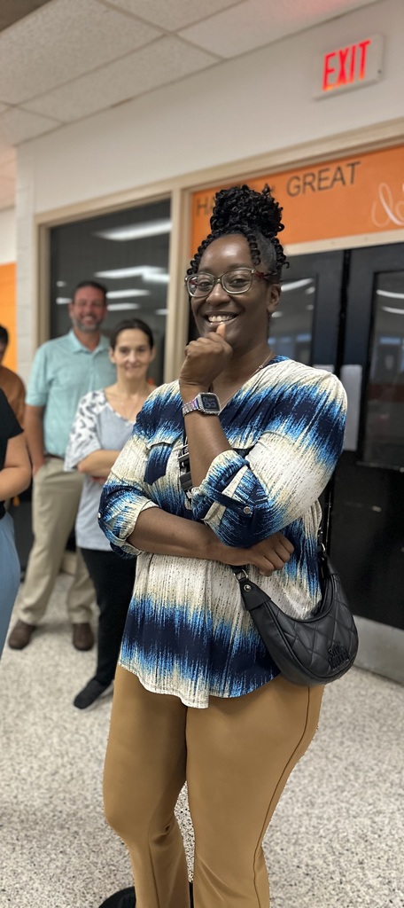 Woman in blue patterned blouse smiles with hand on chin, standing in front of two blurred people.