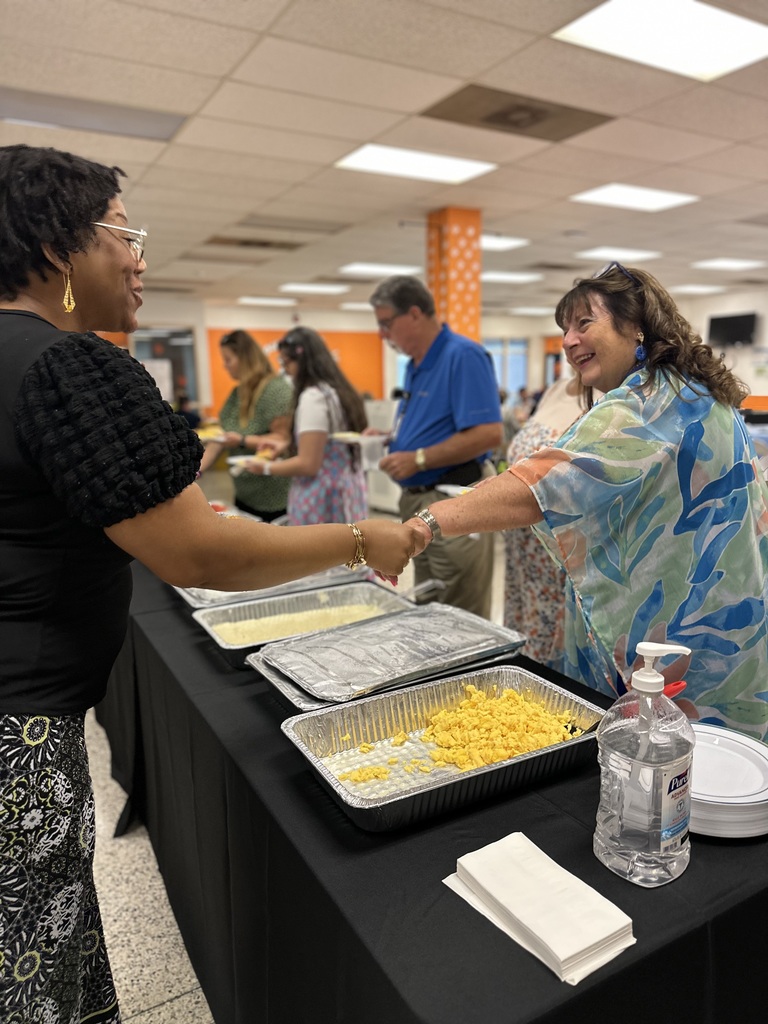 Two women greet and shake hands at a buffet line with trays of scrambled eggs in a cafeteria.