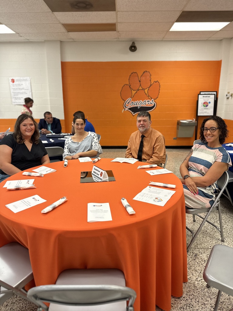 Four people sit at an orange table with Cougar paw wall art in a school cafeteria