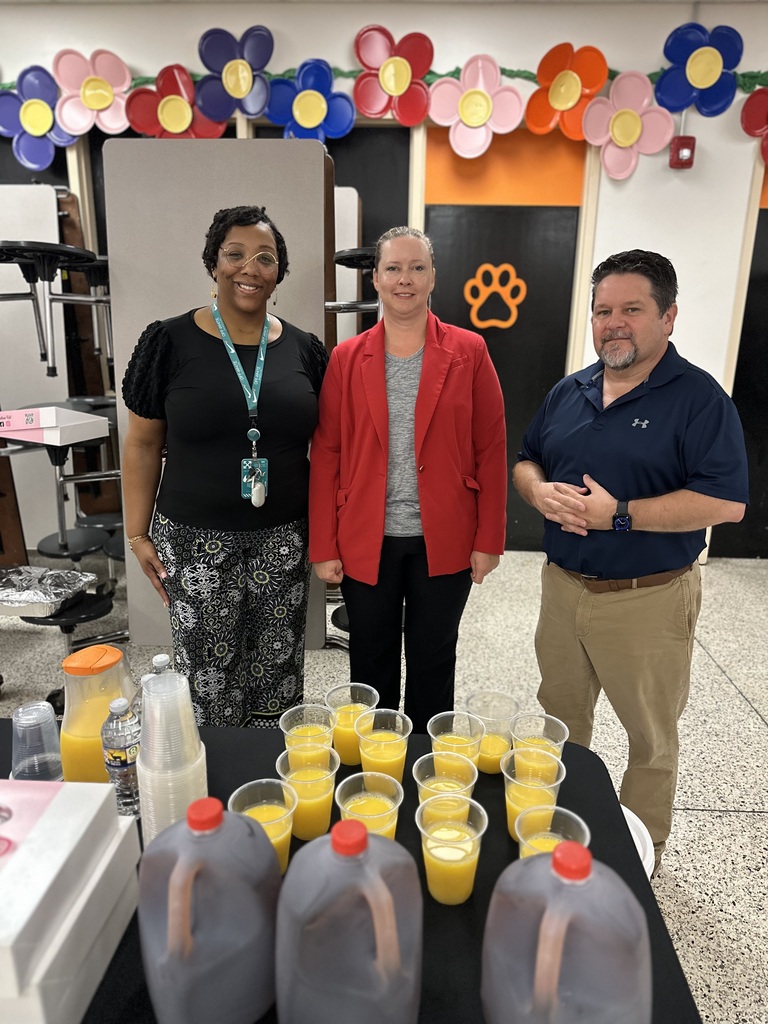Three people stand behind a table with orange juice cups and tea jugs in a decorated cafeteria