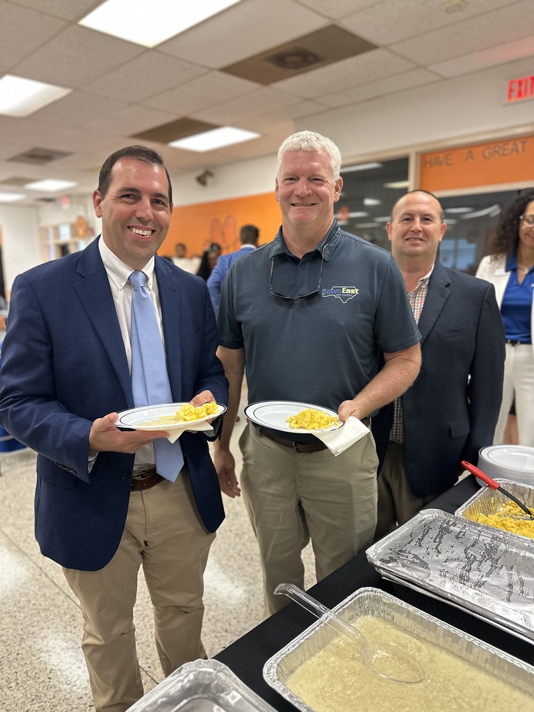 Three men holding plates of scrambled eggs stand by buffet trays in a cafeteria.