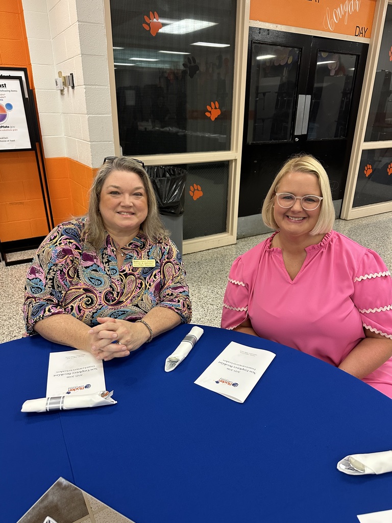 Two women smile at a blue table with folded napkins and programs in front of them.