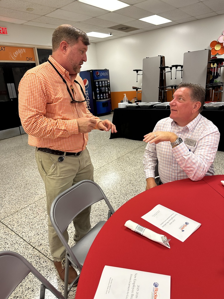 Two men in orange shirts talk at a red table in a cafeteria with folded tables in the background