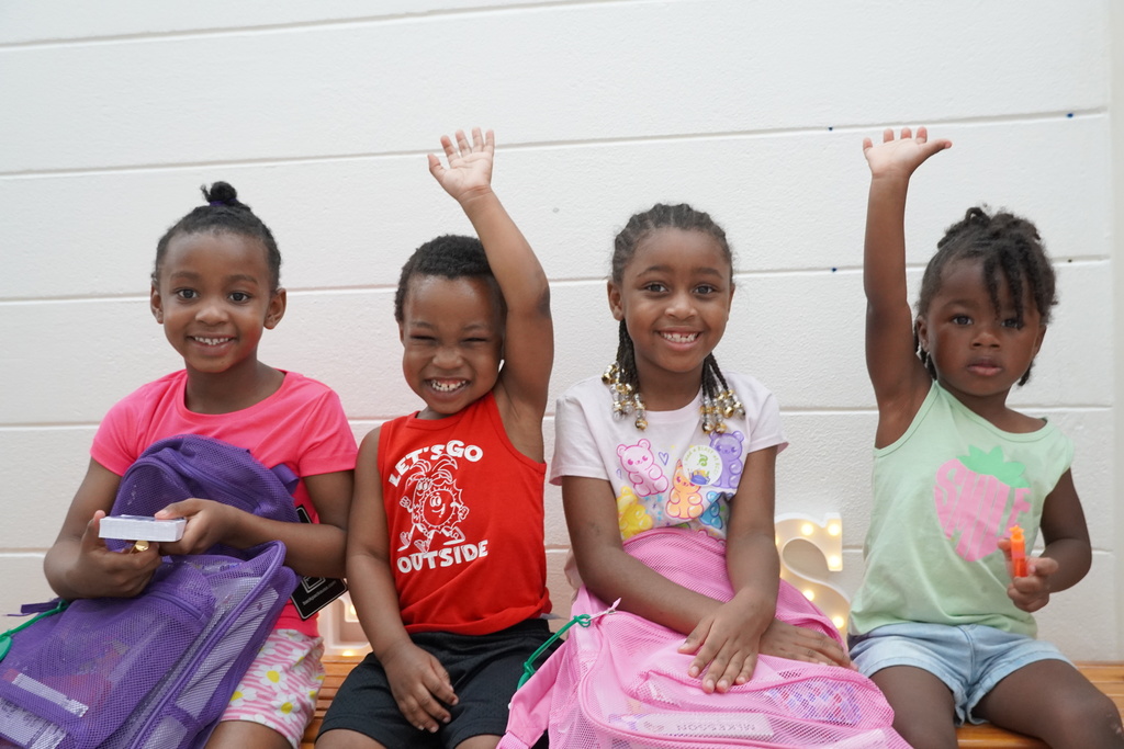 Four young children with backpacks smiling and raising hands while seated on bench