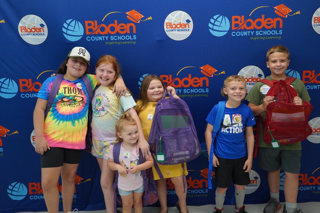 Six children smiling with backpacks posing together in front of district backdrop