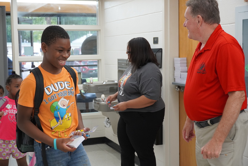 Student smiles while entering school with backpack, greeted by staff