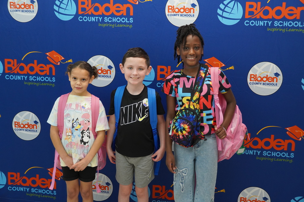Three elementary students with backpacks smiling in front of Bladen County Schools backdrop