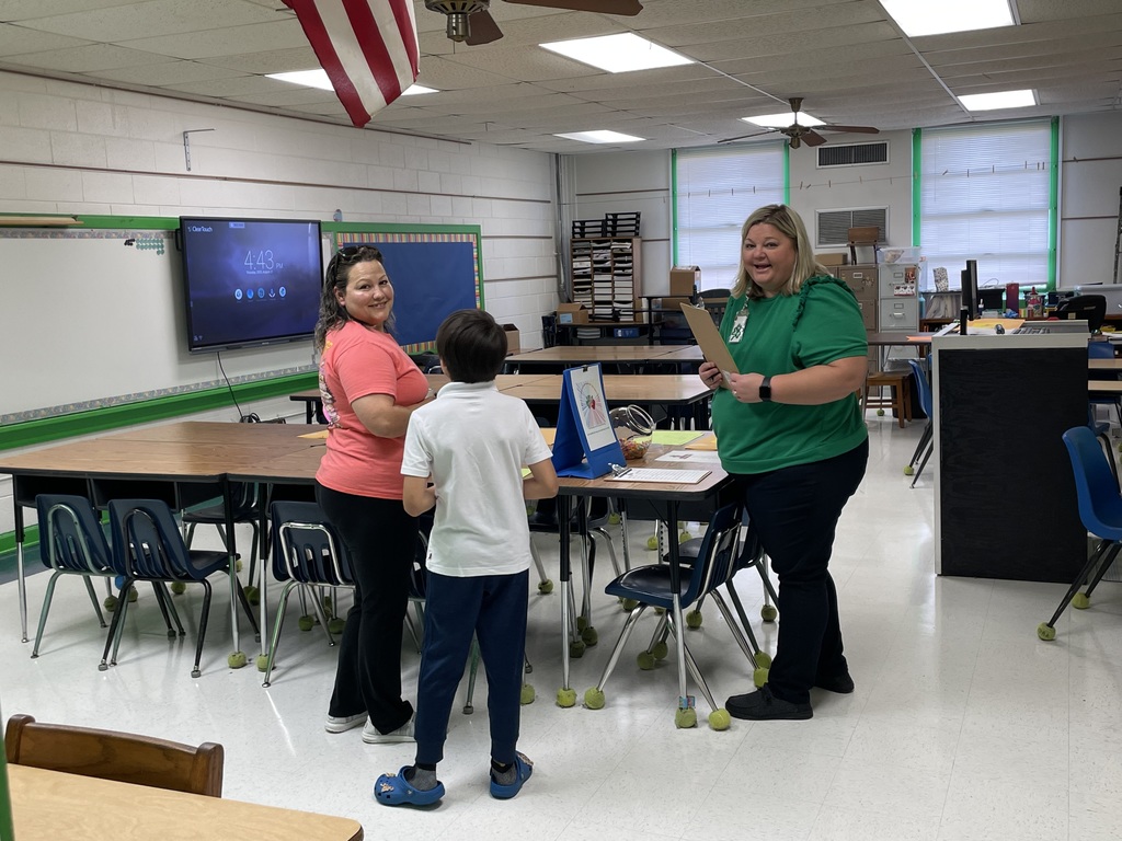 Teacher welcomes a student and parent in a classroom during Open House, smiling with a clipboard
