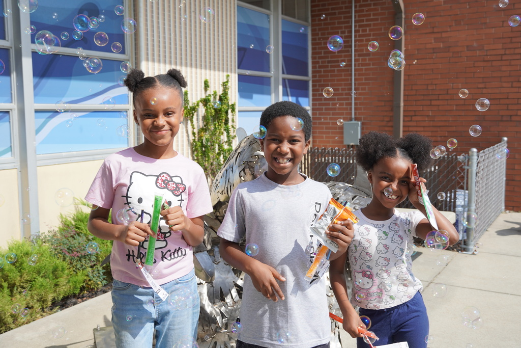 Three smiling children hold treats and stand among floating bubbles outside during Open House.