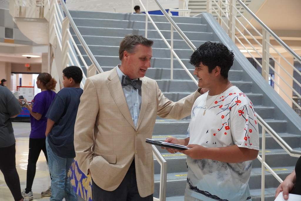 Principal talks with a smiling student holding a folder in the school lobby.