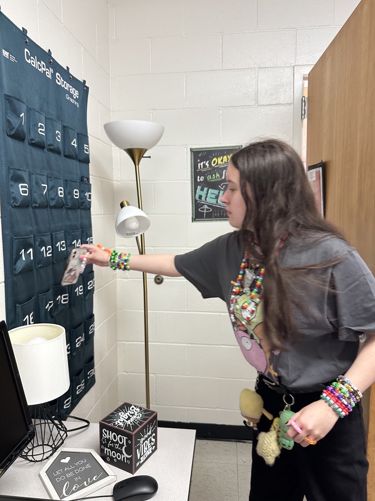 A student places a cell phone into a numbered pocket on a wall organizer in a classroom, supporting a distraction-free learning environment.