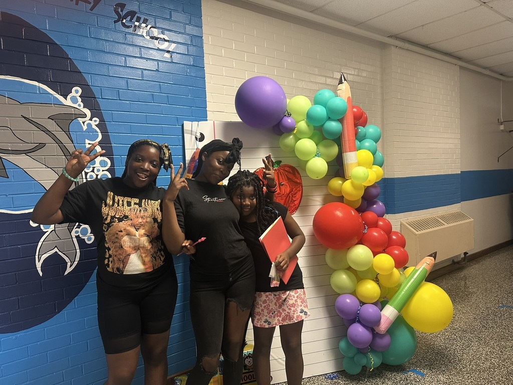 Three students smiling and posing in front of a colorful balloon display with pencils.