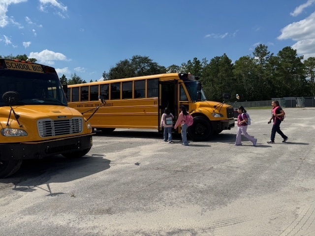 A group of students with backpacks are boarding a yellow school bus parked in a lot on a sunny day. Another school bus is parked nearby. There are trees in the background and the sky is mostly clear with a few clouds.
