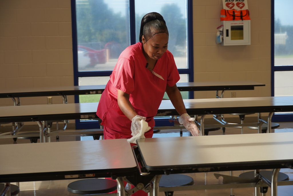 Staff member in red uniform wipes down cafeteria tables to prepare for students.