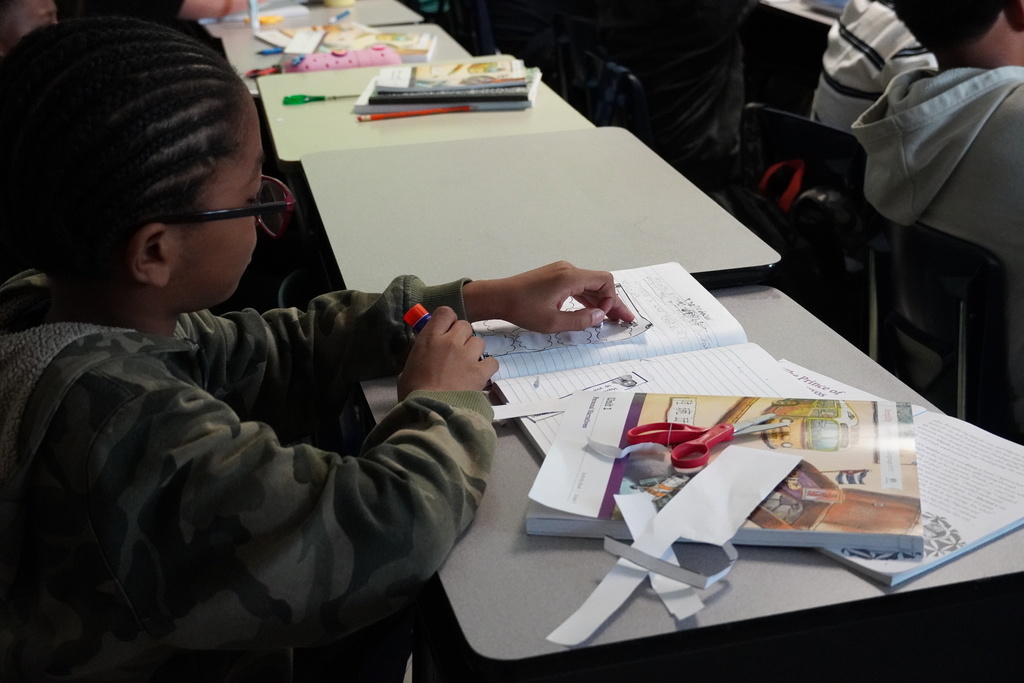 Student works at desk cutting and gluing paper into a notebook during class