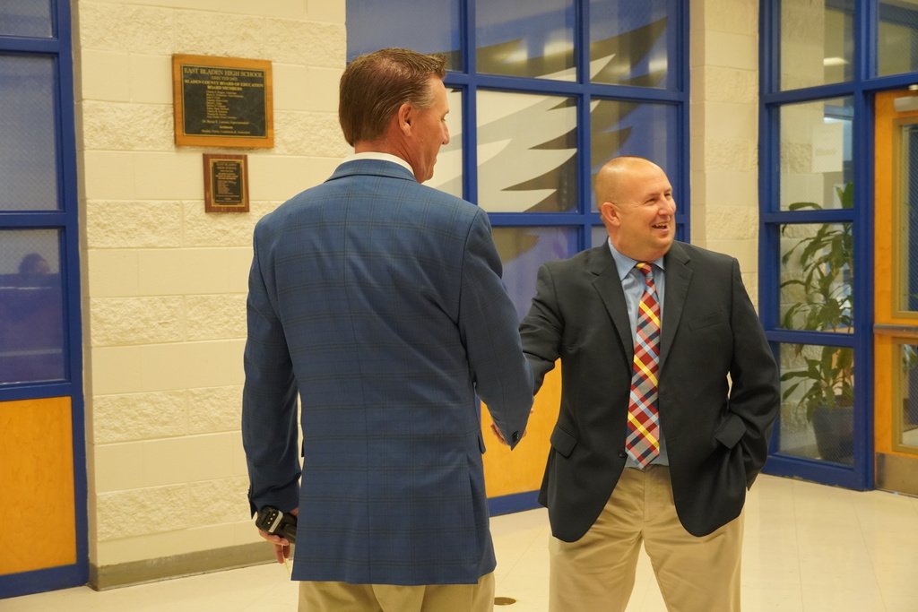 Two men in suits shake hands and smile in East Bladen High School hallway.