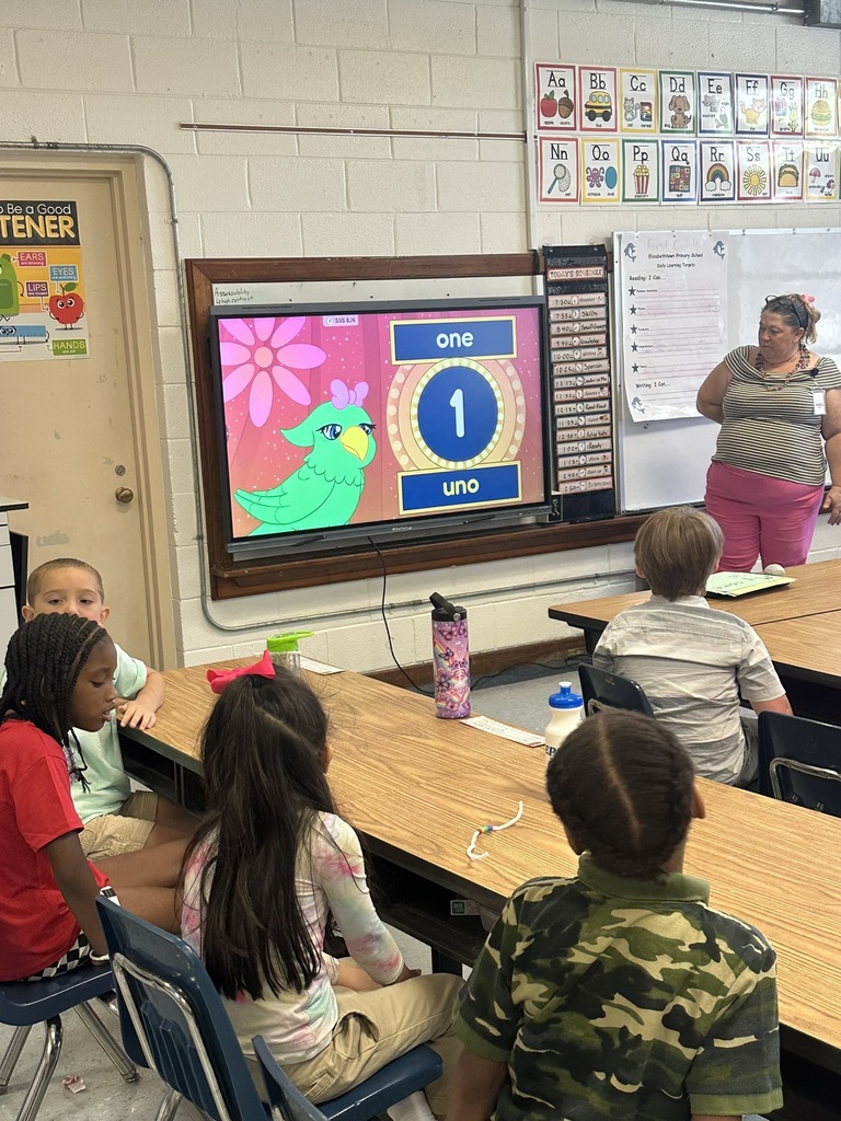A teacher leads a lesson with a bright screen showing “one/uno” while students sit at tables watching.